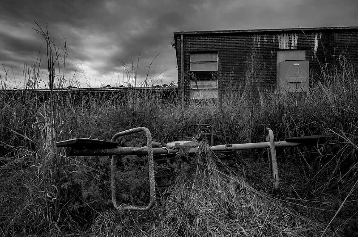 Photo of a rusting see-saw in an overgrown playground. Image converted to monochrome from the original.